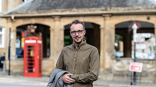 Adam Dance MP stands in the street, smiling at the camera. He is wearing a green button-up shirt and holding a grey coat. Behind him is a historic stone building with arches and a traditional red telephone box. The scene appears to be in a town centre with soft, natural lighting.