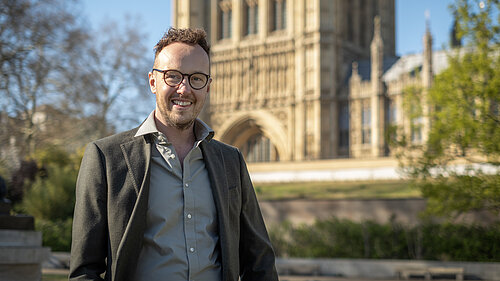 A man wearing glasses stands smiling outdoors in front of a large historic building.
