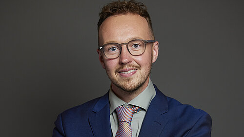 Adam Dance MP smiling, wearing a blue suit, light green shirt, and patterned tie, standing against a plain dark background with arms crossed.