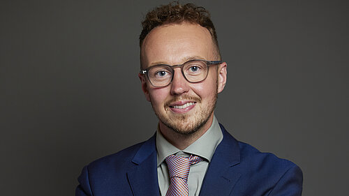 Adam Dance MP standing against a plain dark background, smiling with arms folded. He is wearing glasses, a blue suit, a light green shirt and a red and blue patterned tie.