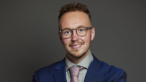 A man in a blue suit, light shirt and patterned tie smiles at the camera against a plain dark background.