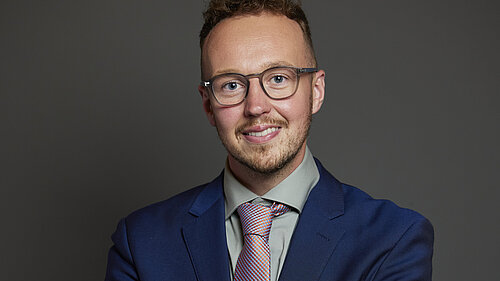 Adam Dance MP poses for a formal portrait against a plain dark background. He is smiling and wearing a navy blue suit, light grey shirt, and patterned tie. He is wearing glasses.
