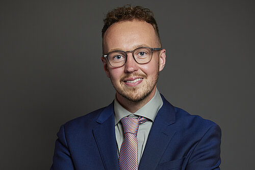 Adam Dance MP poses for a formal portrait against a plain dark background. He is smiling and wearing a navy blue suit, light grey shirt, and patterned tie. He is wearing glasses.