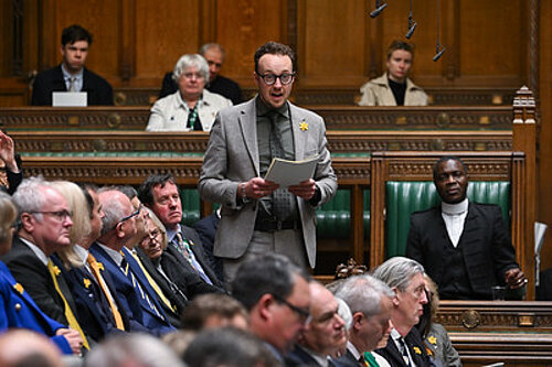 A man stands speaking and holding papers in the House of Commons chamber.