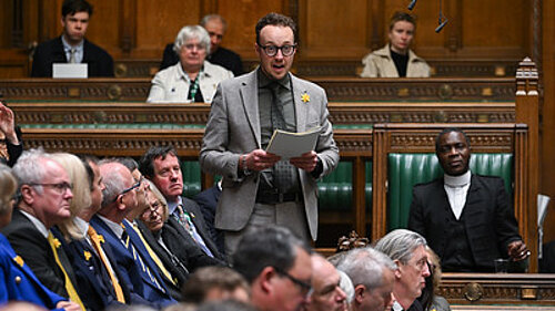 A man stands speaking and holding papers in the House of Commons chamber.