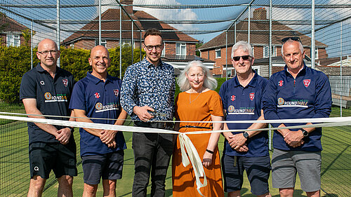 Steve Redwood, Scott Ewens, Adam Dance MP, Trudy Dyke, Rob Fox (Chairman), and Richard Reeves (President of Yeovil Cricket Club) stand together in a cricket training net area on a sunny day. Adam is holding scissors after cutting a ceremonial ribbon to mark the opening of a new cricket facility, with everyone smiling towards the camera.