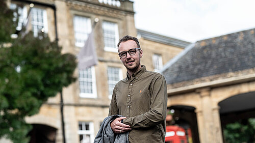 Adam Dance MP standing outside in front of a historic stone building, holding a grey coat and smiling at the camera.