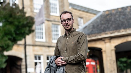 Adam Dance MP stands outside in a town centre, smiling at the camera. He is wearing a green button-up shirt and holding a grey coat. Behind him is a historic stone building and a traditional red phone box. The scene is bright with natural daylight.