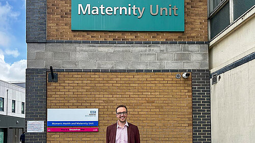 A man stands outside a hospital building under a sign for the maternity unit.