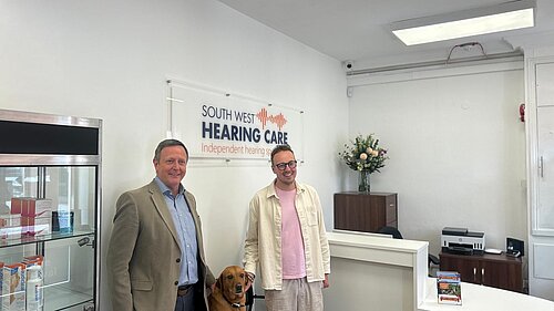 Two men and a guide dog stand in the reception area of South West Hearing Care beneath the company sign.