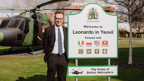 A man stands beside a sign reading “Welcome to Leonardo in Yeovil” with a helicopter displayed behind him.