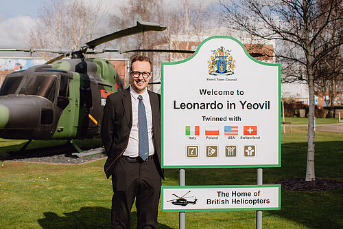 A man stands beside a sign reading “Welcome to Leonardo in Yeovil” with a helicopter displayed behind him.
