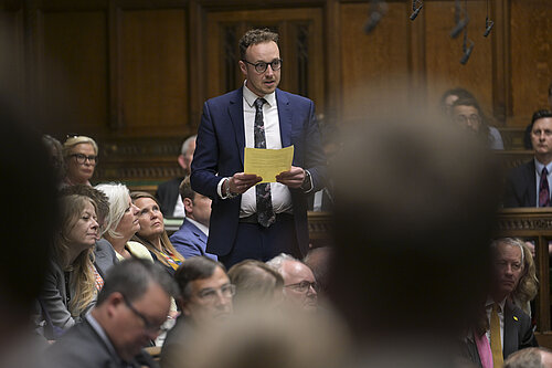 Adam Dance MP stands and speaks in the House of Commons, holding a sheet of yellow paper. He is wearing a blue suit and floral tie. Other MPs are seated around him, listening attentively.