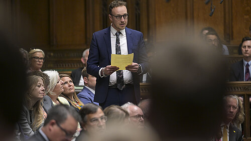 Adam Dance MP stands and speaks in the House of Commons, holding a sheet of yellow paper. He is wearing a blue suit and floral tie. Other MPs are seated around him, listening attentively.