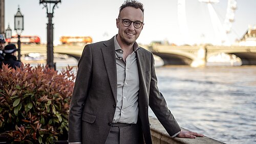 Adam Dance MP stands smiling by the River Thames in London. He is wearing a grey suit and open-collar shirt. The London Eye and Westminster Bridge are visible in the background on a bright day.