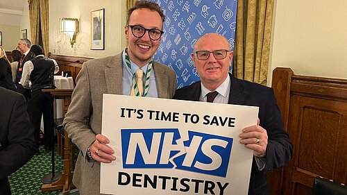 Two men stand indoors smiling at the camera while holding a sign that reads It’s time to save NHS dentistry.