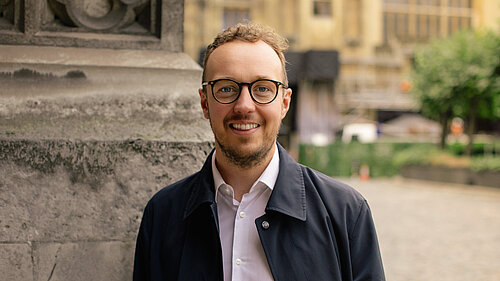 Adam Dance MP smiling in front of a stone pillar outside the Houses of Parliament, wearing glasses, a white shirt, and a dark jacket.