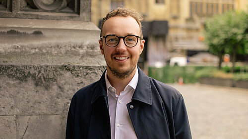 A man stands in a shirt and a black coat, wearing glasses with a historic building in the background.