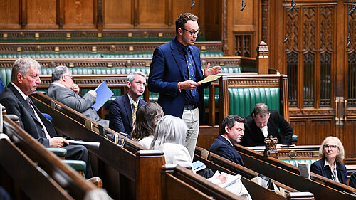 Adam Dance MP stands while others sit in a parliamentary chamber with green leather seats and wooden paneling.