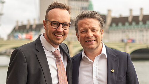 Adam Dance MP and Jamie Oliver stand side by side, smiling, with the River Thames and Westminster buildings in the background. Both are dressed in suits, with Adam wearing a pink tie and Jamie in an open-collared white shirt and dark blazer. The London Eye is partially visible on the left under a cloudy sky.