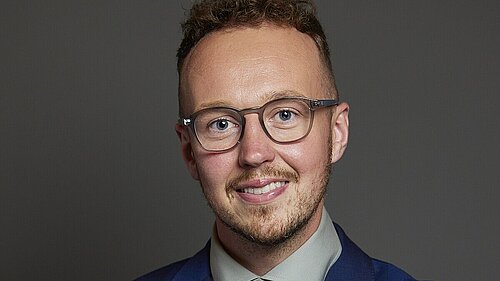 Adam Dance MP standing against a plain dark background, smiling with arms folded. He is wearing glasses, a blue suit, a light green shirt and a red and blue patterned tie.