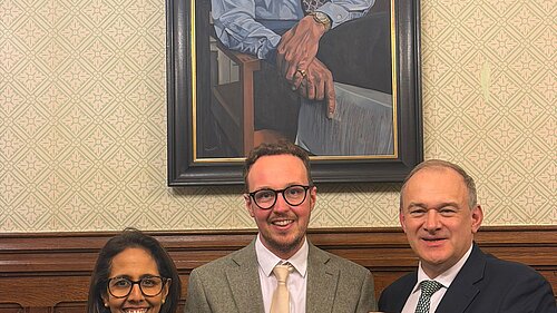 Adam Dance MP stands with two colleagues in Parliament holding a printed Bill, with a large painted portrait hanging on the wall behind them.