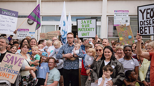 Adam Dance MP stands holding a baby among a large group of parents, children, and supporters outside a hospital. They are holding handmade signs that say things like "Save Our Maternity Unit" and "Yeovil Deserves Better," as part of a protest to protect local maternity services.