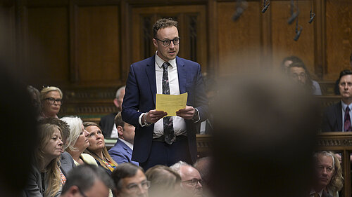 Adam Dance MP stands in the House of Commons, surrounded by MPs. He wears a blue suit and tie, a white shirt, and holds a small piece of paper.