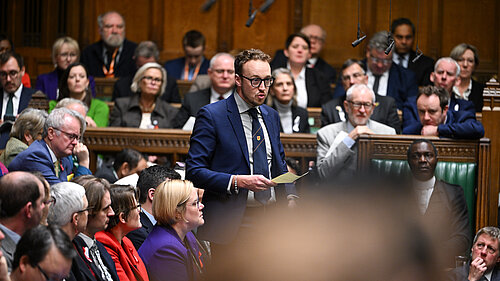 A man in a suit stands speaking in the House of Commons chamber, holding a piece of paper. Other MPs sit and listen around him.