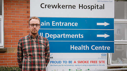 Adam Dance MP standing in front of a sign for Crewkerne Hospital in Somerset.