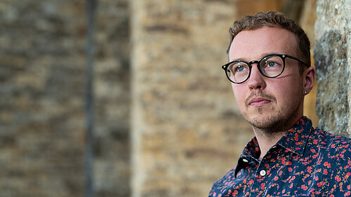 Adam Dance MP standing beside a stone wall, wearing glasses and a dark shirt with a red floral pattern, looking thoughtfully into the distance.