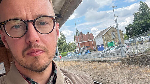 A man wearing glasses and a beige jacket standing on a train platform, with railway tracks and buildings behind him.