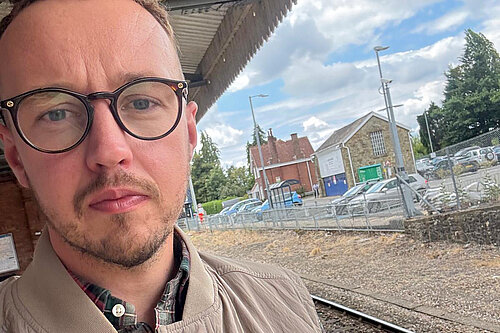 A man wearing glasses and a beige jacket standing on a train platform, with railway tracks and buildings behind him.