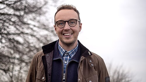 Adam Dance MP smiling outdoors on a cloudy day, wearing glasses, a checked shirt, navy jumper, and brown Barbour jacket, with bare winter trees in the background.