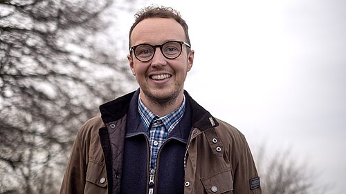 Portrait of a man wearing glasses and a brown coat, with a tree in the background. 