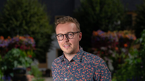 Adam Dance MP standing outdoors in a garden with colourful flowers and greenery, wearing a dark shirt with a red floral pattern and glasses.