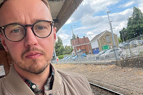 "A man in glasses and a beige jacket takes a selfie on a train platform, with the tracks, station building, and car park visible behind him under a partly cloudy sky."