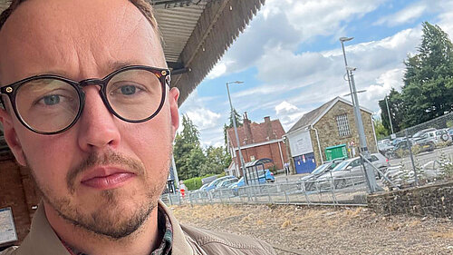 "A man in glasses and a beige jacket takes a selfie on a train platform, with the tracks, station building, and car park visible behind him under a partly cloudy sky."
