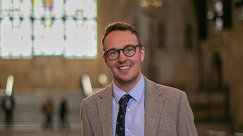 Adam Dance MP stands smiling inside Westminster Hall, part of the Houses of Parliament. He wears a light grey suit, blue shirt, and navy tie with a subtle pattern. Behind him, the grand arched window and stone staircase are softly out of focus, highlighting the historic setting.