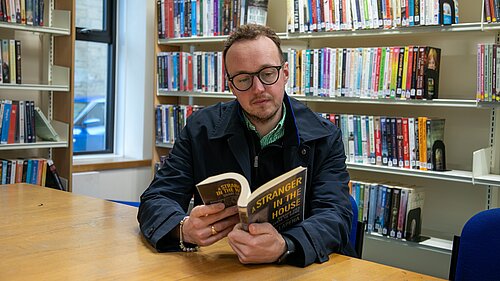 A man sits at a table in a library reading a book, with shelves of books behind him.