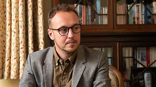 Adam Dance MP seated indoors, wearing a grey blazer and brown shirt, looking thoughtfully to the side. A bookcase and patterned curtains are in the background.