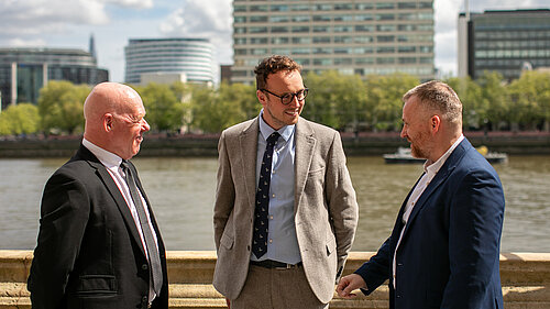 Peter Brown (Technical & Development Director at the CPA), Adam Dance MP, and Steve Mulholland (CPA Chief Executive) stand together outside by the River Thames, engaged in conversation, with tall buildings and trees in the background.