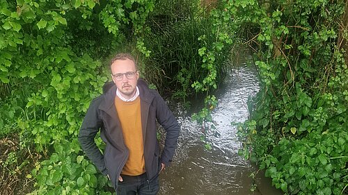 Adam Dance MP stands beside a small stream surrounded by lush green vegetation, looking up toward the camera.