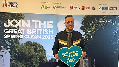 Adam Dance MP smiling while holding a heart-shaped sign that reads “Love Where You Live – Keep Britain Tidy” in front of a display for the Great British Spring Clean 2025.