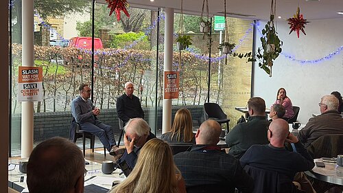 A group of people sit in a café style room listening to two men speaking at the front. Posters on the windows read Slash VAT for Hospitality and Save Our High Streets. Christmas decorations and hanging plants are visible.