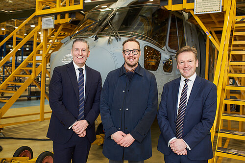 Three men stand smiling in front of a helicopter inside a factory, with yellow metal stairs and platforms around them.