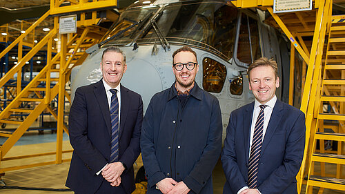 Three men stand smiling in front of a helicopter inside a factory, with yellow metal stairs and platforms around them.